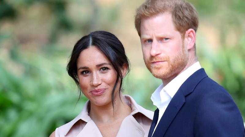 Meghan Markle, in a khaki dress, stands with Prince Harry, in a blue suit, outdoors