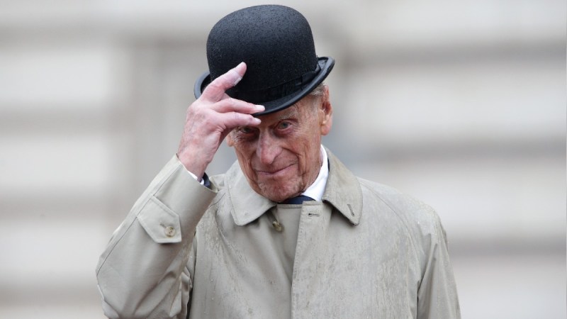 Prince Philip tips a black bowler hat while wearing a tan jacket outdoors