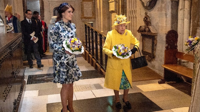 Princess Eugenie, in a blue and white dress, holds flowers in her hands and stands with Queen Elizabeth, dressed in yellow, who holds a similar bouquet
