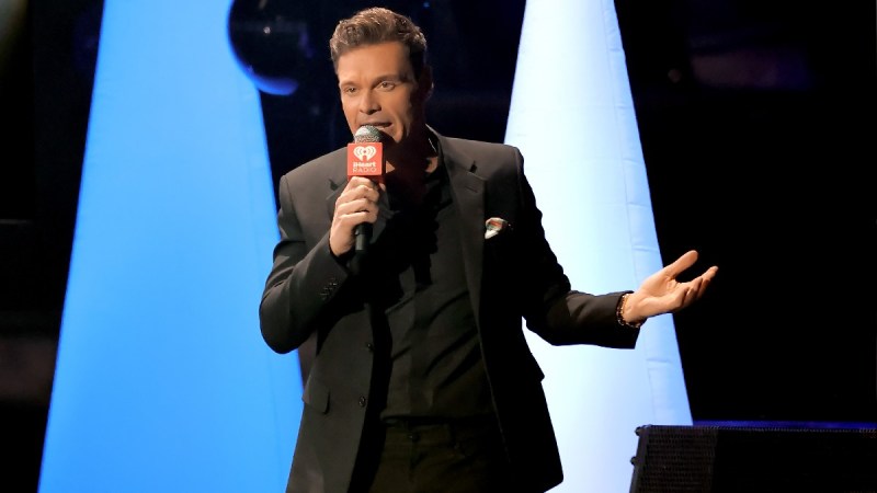 Ryan Seacrest wears a black suit onstage as he hosts an event