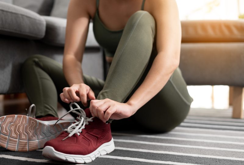 Woman sitting on floor of home lacing up workout shoes.