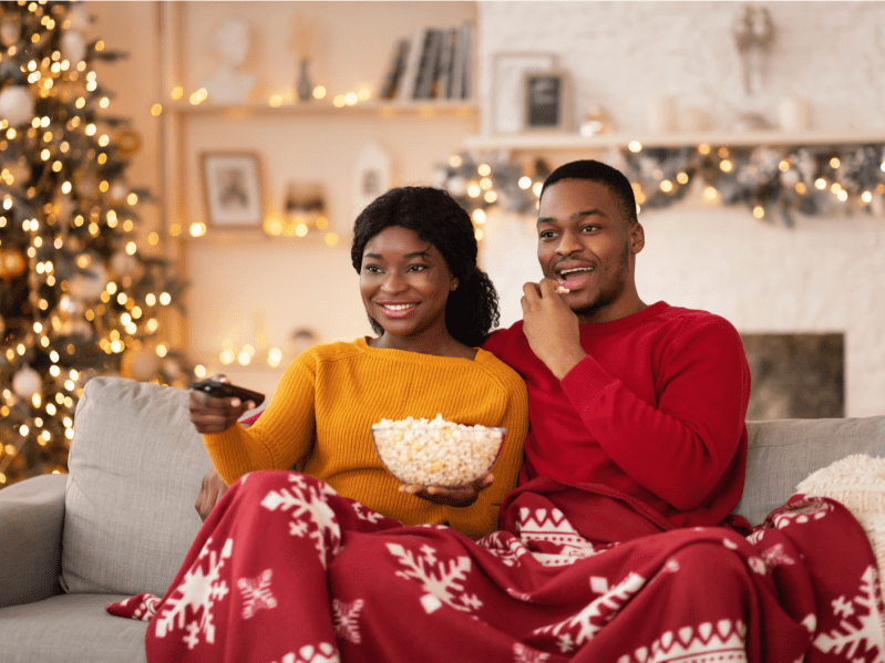 Free time, favorite movie and self-isolation at home. Millennial funny african american husband and wife with remote control and popcorn covered with blanket and watching tv in interior with tree