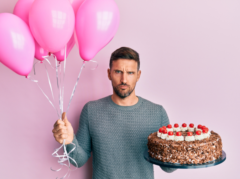Angry man holding birthday cake and balloons on a pink background