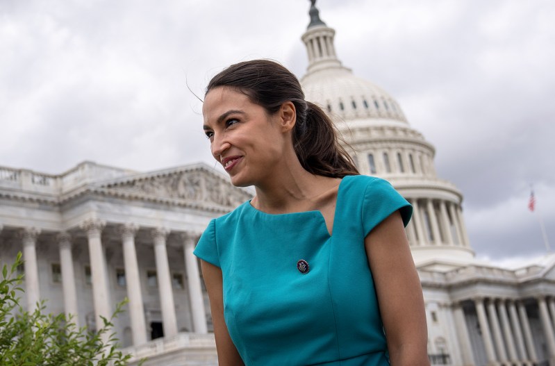 A smiling Alexandria Ocasio-Cortez in a green dress, standing in front of the Capitol.