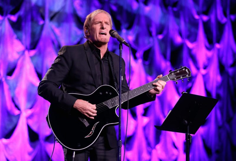 Michael Bolton singing, holding a black guitar, with a pink/purple background