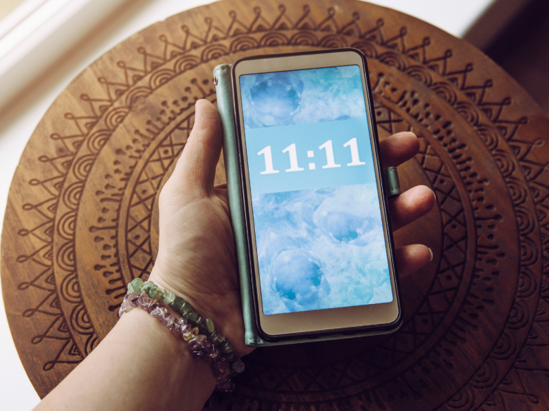 Arm with bracelets holding smart phone with cloud background and 11:11 time stamp, circular mandala mat in the background