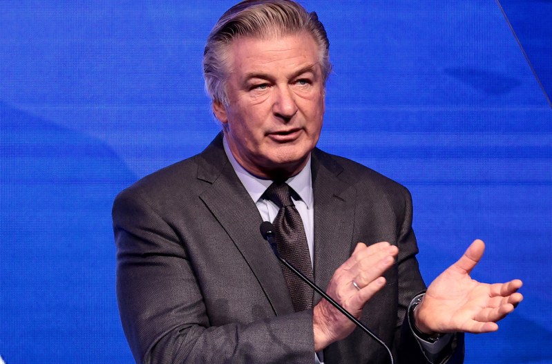 Alec Baldwin in a grey suit speaking at a podium