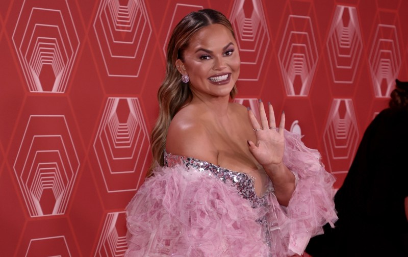 Chrissy Teigen wears a silver and pink dress against a red background on the red carpet