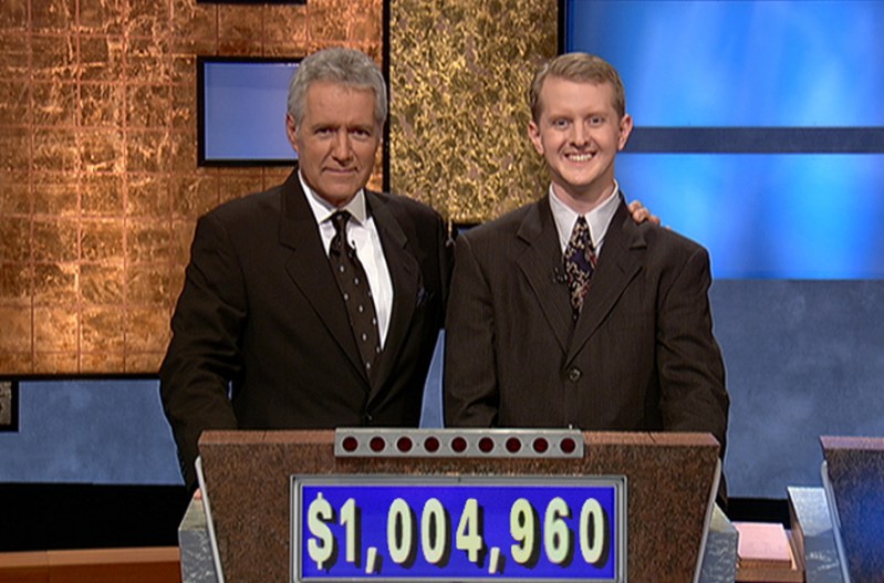 Alex Trebeck standing with Ken Jennings behind a contestant podium on Jeopardy!