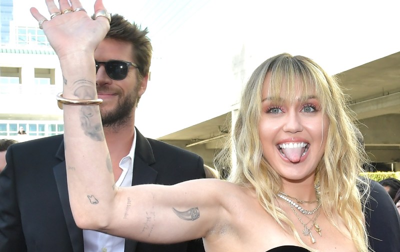 Miley Cyrus, in a black dress, waves while walking with Liam Hemsworth, in a black suit, on the red carpet