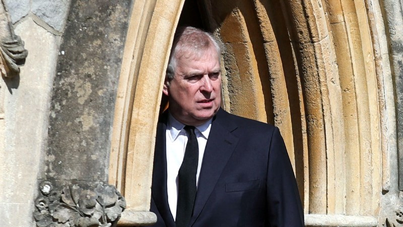 Prince Andrew wears a dark suit and peers out from a doorway at a chapel in Windsor