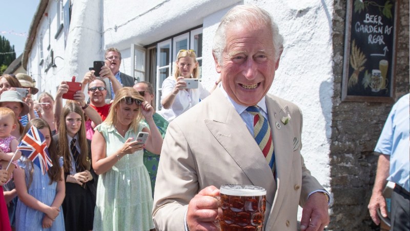 Prince Charles holds up a glass mug of beer while wearing a khaki suit outdoors during a royal visit