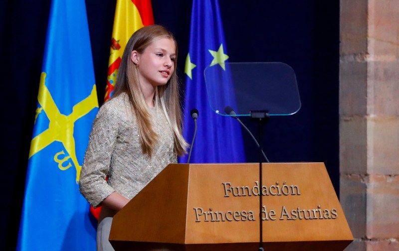 Princess Leonor wears a beige dress and stands behind a wooden podium as she makes remarks