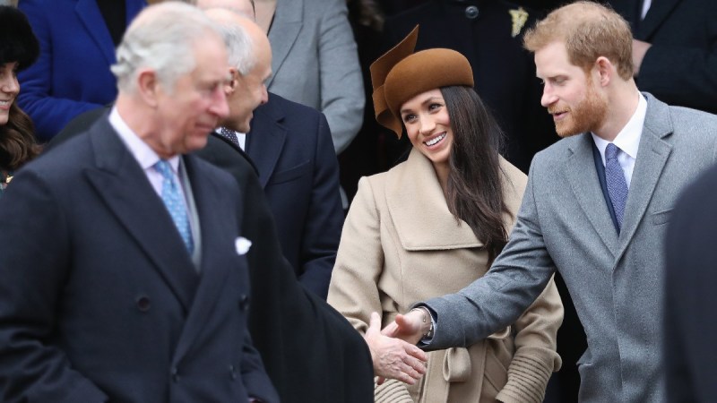 Prince Charles walks by in the foreground as Meghan Markel and Prince Harry greet someone in the background