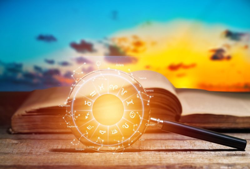 Magnifying glass with astrology diagram in the lens in front of open-faced book and sunset background