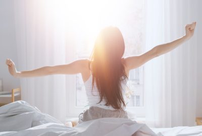Woman stretching arms out while sitting on a bed in a sunlight room.