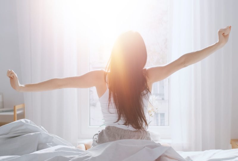 Woman stretching arms out while sitting on a bed in a sunlight room.