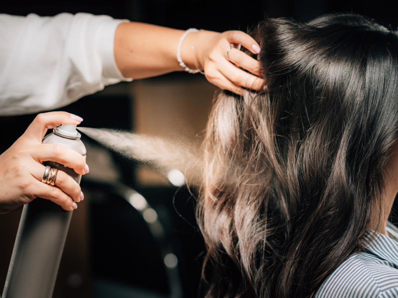 Hairdresser spraying woman’s long black hair with hair spray.