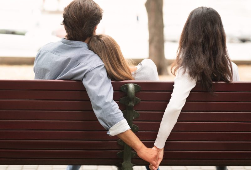 Back view of three people sitting on bench, first and third person holding hands behind middle person, who is resting head on first person's shoulder