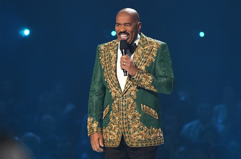 Steve Harvey smiling on stage with a microphone wearing a very loud gold jacket