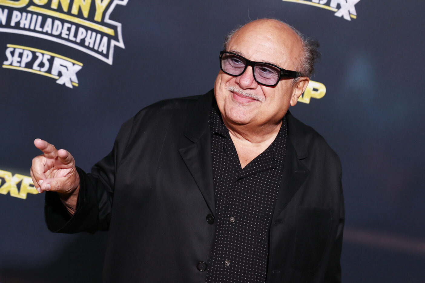 Danny DeVito smiling at a camera, wearing a black dress shirt