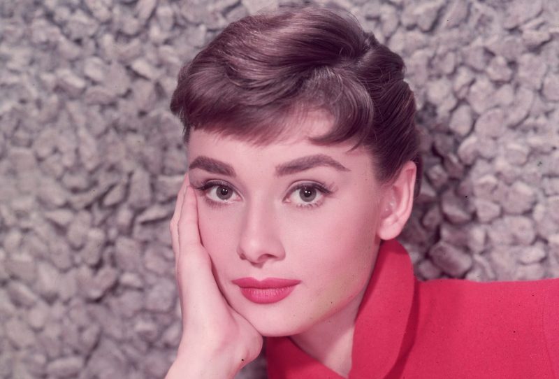 Headshot portrait of Belgian-born actor Audrey Hepburn (1929 - 1993) leaning on her hand in a red jacket with a poodle applique.