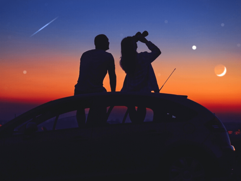Silhouette of couple sitting on car and using binoculars to look out at colorful starry sky