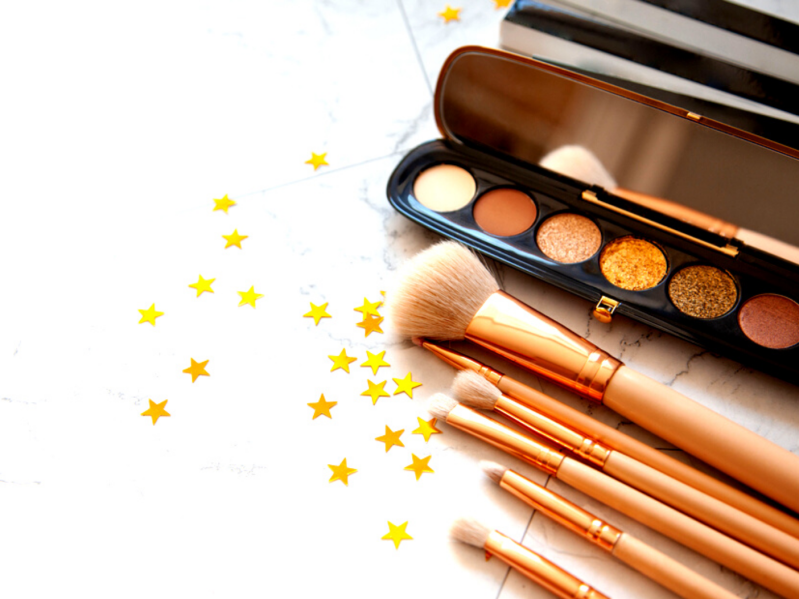 Cosmetic brushes and palette on white table surrounded by gold star confetti