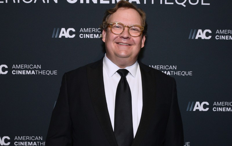 Andy Richter wears a black suit against a black background on the red carpet