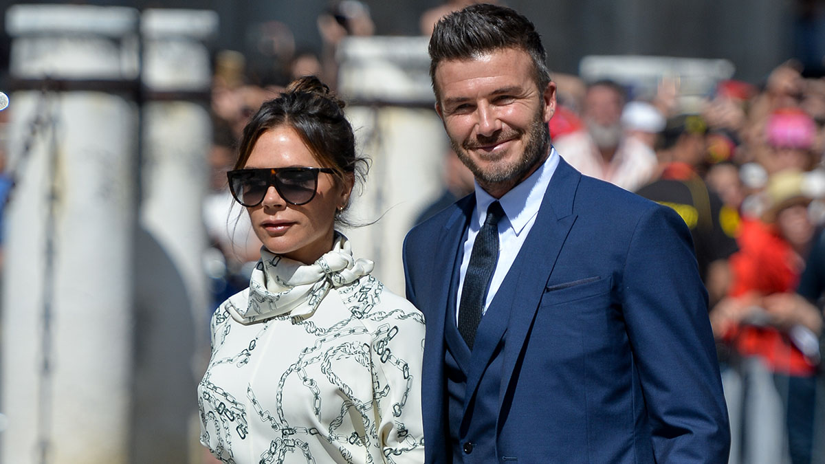 Victoria Beckham in a white dress, wearing sunglasses walking with David Beckham in a suit, smiling