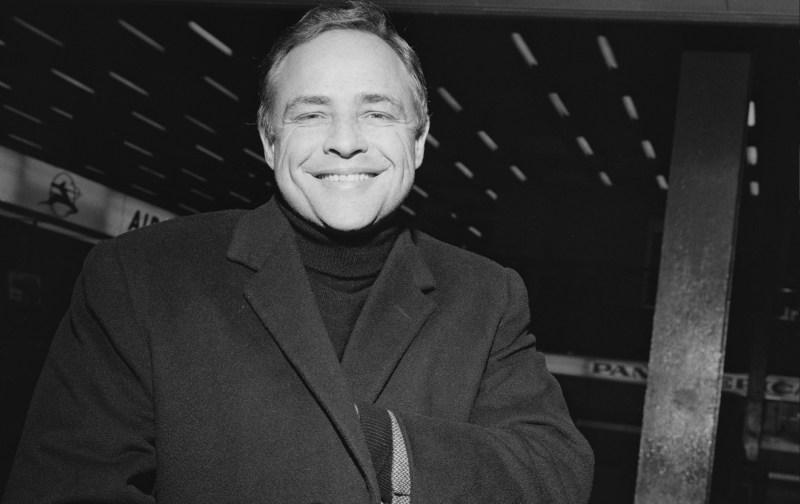 Marlon Brando smiles in a black and white photo while wearing a dark colored coat