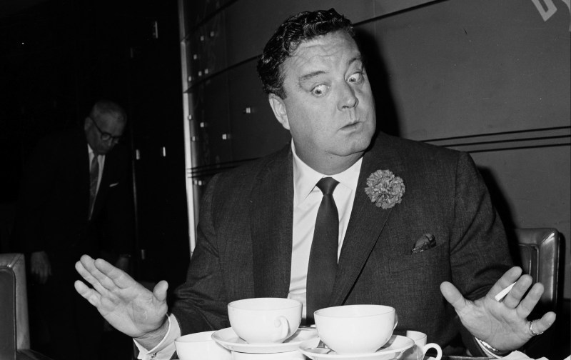 Jackie Gleason pulls a comedic face while seated at a table and wearing a dark suit
