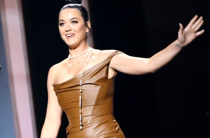 Katy Perry in a brown dress with her arm out to her left.