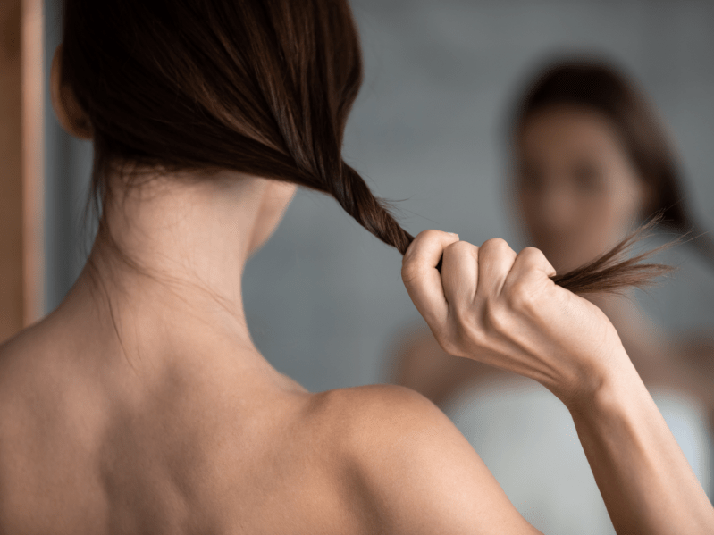 Rear view close up woman holding long straight thin hair in hand, checking split dry ends, young female with naked shoulders standing in front of mirror in bathroom, hairstyle and care
