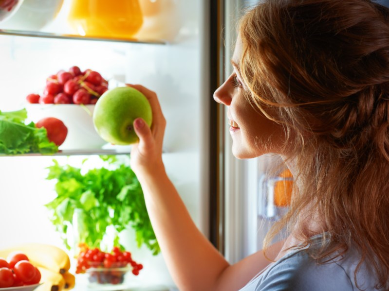 Woman in the dark at the open refrigerator and removing apple from fridge