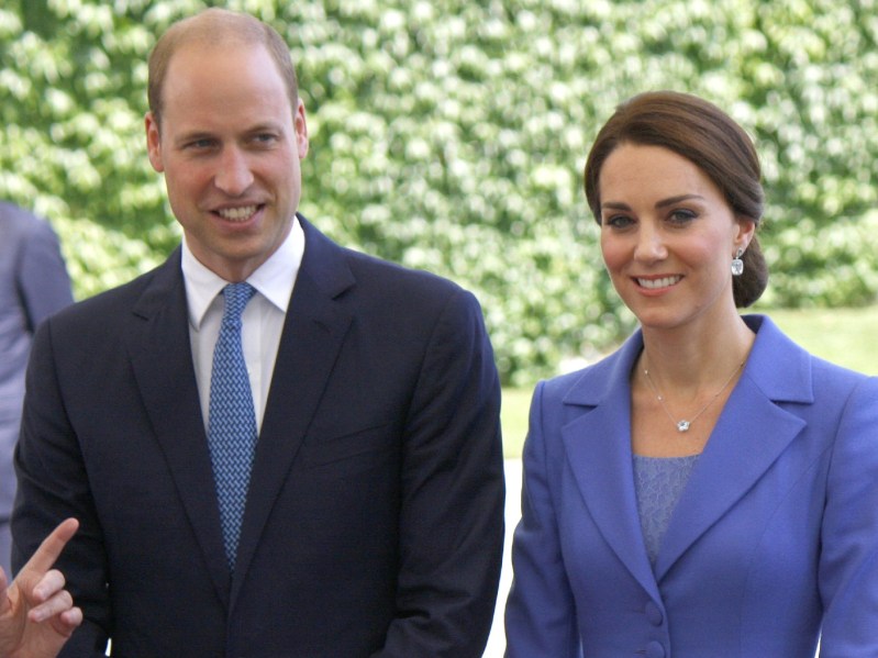 Prince William wearing black suit with blue tie standing. next to Kate Middleton wearing powder blue suit jacket