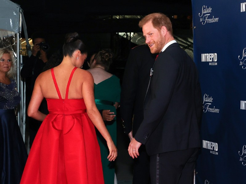 Prince Harry and Meghan Markle, in a black suit and red dress respectively, walk a red carpet at a Veterans event