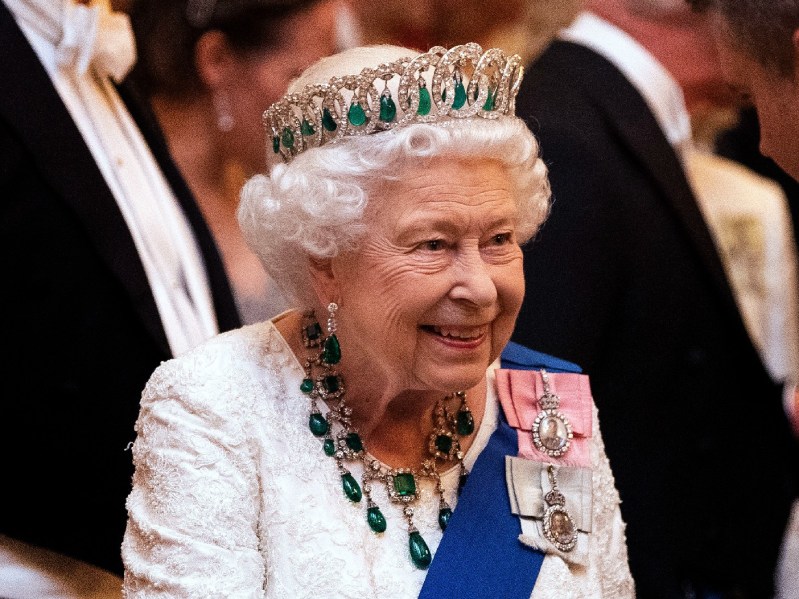Queen Elizabeth wears a white gown with a blue sash and emerald tiara and necklace to a royal reception