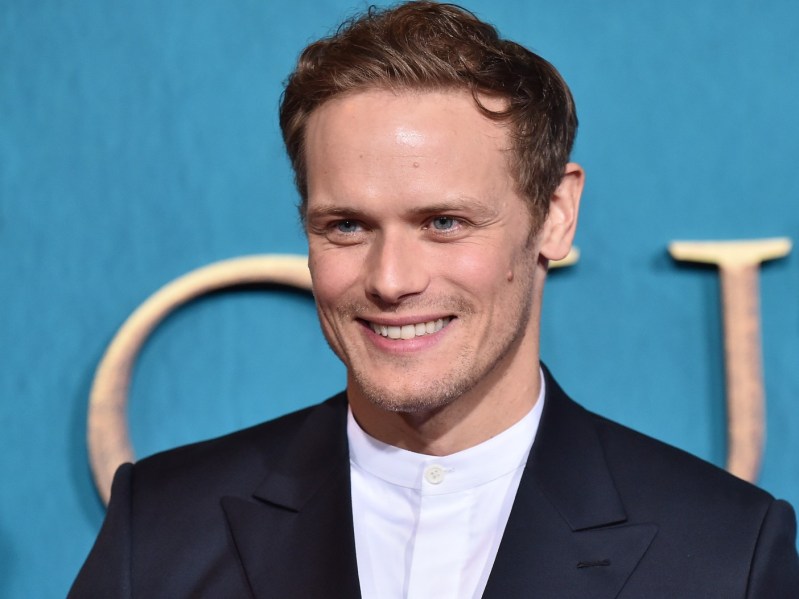 Sam Heughan smiling in a black blazer against a light blue backdrop