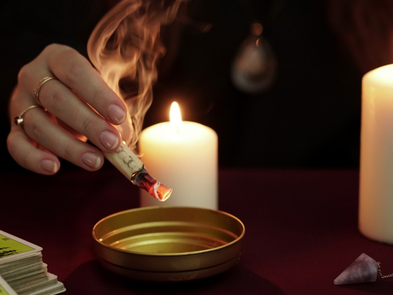 Witch is fortune teller in black mantle holding burning paper with spell over bowl. Tarot cards, amethyst stone, white candles on dark mystic background. Occult, esoteric, divination and wicca concept