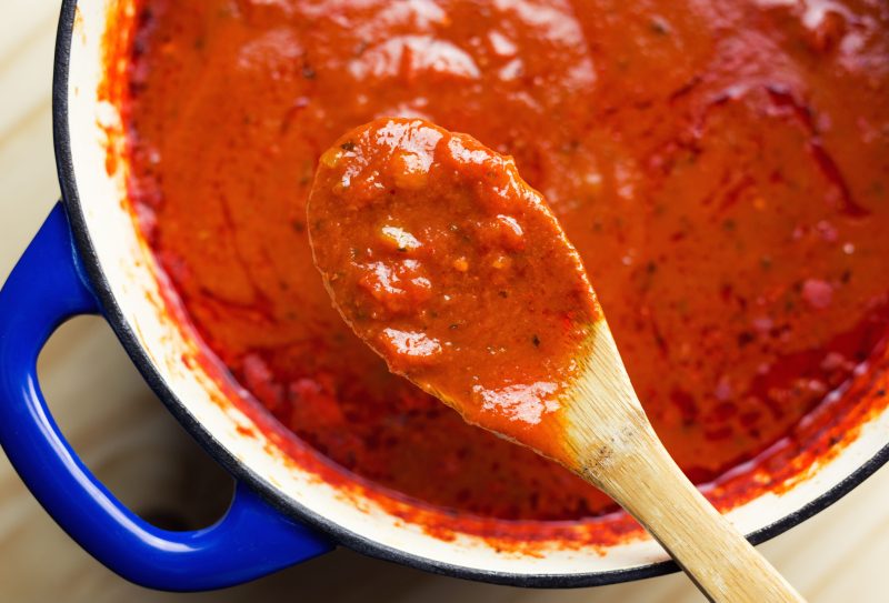 A wooden spoon ladling marinara sauce out of a dutch oven.