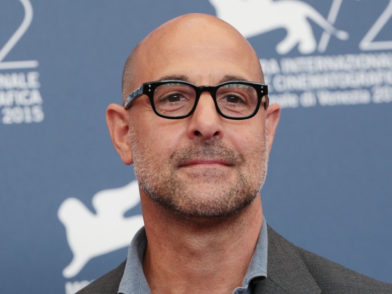 Stanley Tucci smiling wearing glasses against a slate blue backdrop