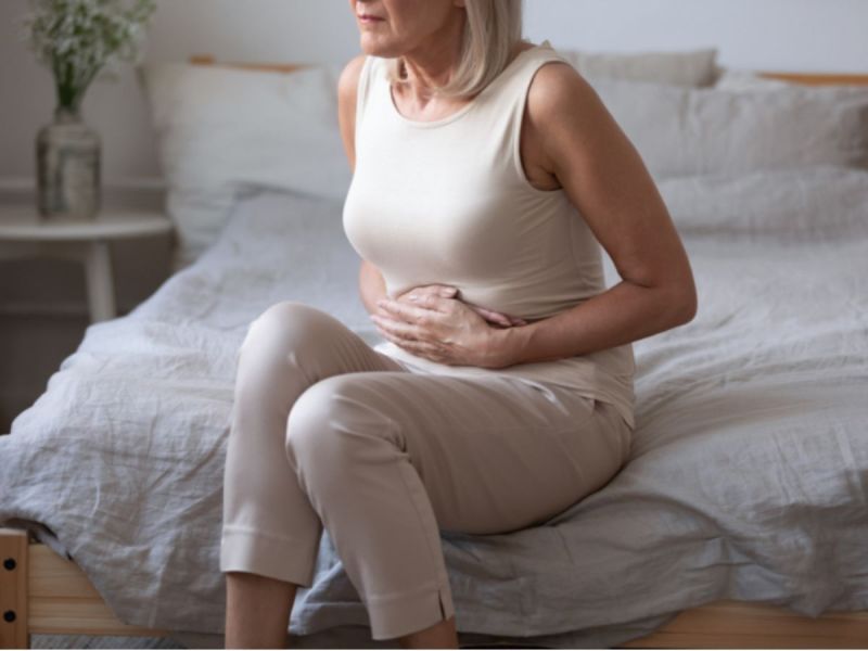 Older woman sits on bed holding stomach