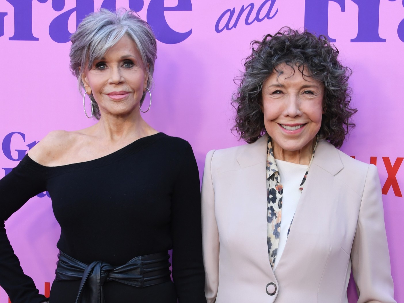 Jane Fonda (L) wearing one-shoulder black dress and Lily Tomlin (R) wearing beige suit jacket