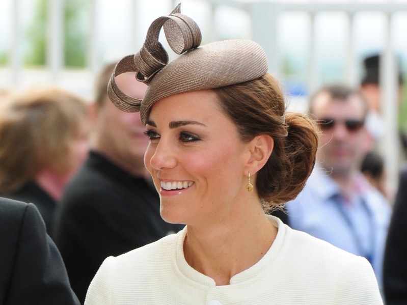 Closeup of Kate Middleton wearing off-white stop and beige hat, smiling off-camera