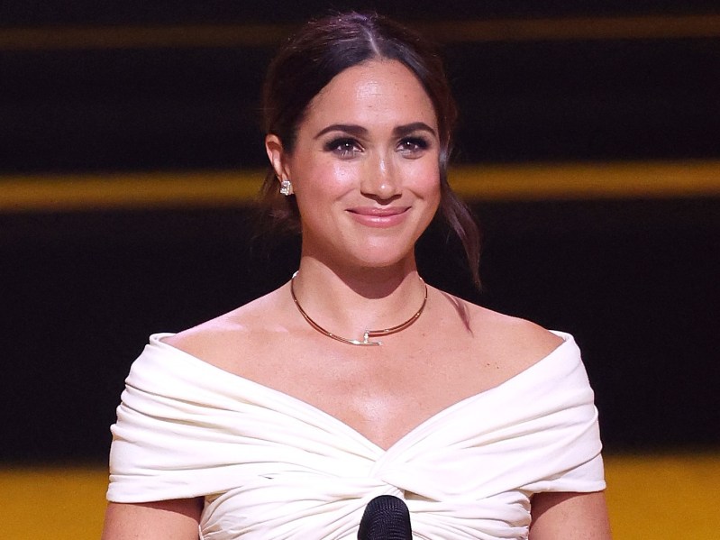 Meghan Markle holding a microphone with off-shoulder white dress and hair pulled back