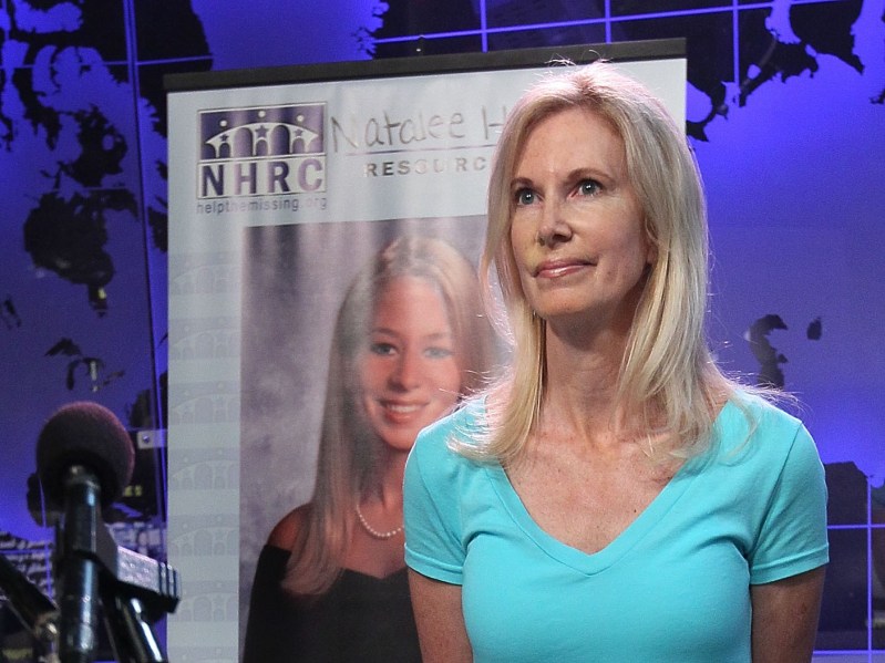 Beth Holloway wearing turquoise shirt, standing in front of missing persons poster for Natalee Holloway