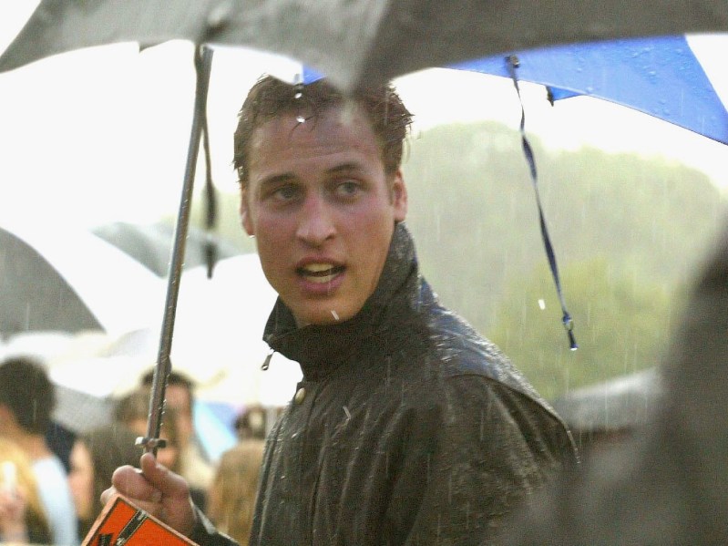 Prince William wears a black jacket and stands beneath an umbrella at a royal sporting event