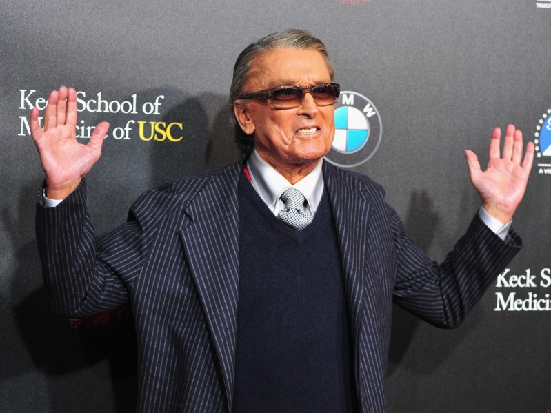 An elderly Robert Evans with both hands up, wearing sweater vest with blazer and sunglasses