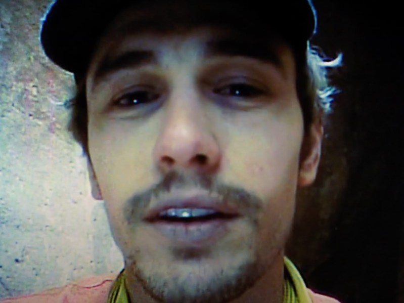 Closeup of James Franco portraying Aron Ralston in the film "127 Hours." the video quality is that of a handheld camcorder. Franco wears a baseball cap and has a slight mustache. He appears to be talking into the camera.